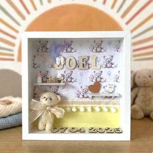 May include: A white shadow box frame containing a personalized display with the name JOEL in wooden letters, a small plush teddy bear with a cream ribbon, miniature shelves, baby-themed icons, and the date 07.04.2025 in wooden numbers.