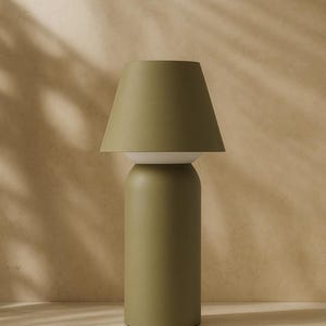 May include: A modern olive green table lamp with a cylindrical base and a conical lampshade. The lamp has a white section between the base and the shade. The background is a neutral beige.