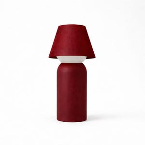 May include: A modern table lamp with a deep red lampshade and base. The lamp has a white midsection, and the shade is a tapered cone shape. The lamp is a solid color and sits on a white background.
