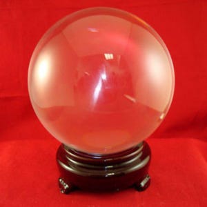 May include: A clear, spherical glass orb resting on a dark brown, circular wooden stand with four small feet. The smooth, transparent surface of the sphere reflects light, creating bright highlights against the solid red background.