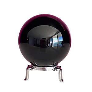 May include: A smooth, polished black sphere resting on a silver-colored metal stand with four curved legs. The surface of the sphere is highly reflective, showing a clear distortion of the surrounding room and a window view.