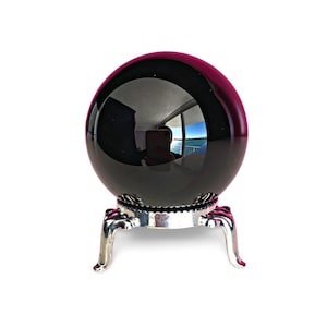 Könnte beinhalten: Eine schwarze Kristallkugel mit reflektierender Oberfläche, die auf einem silberfarbenen Metallständer ruht. Der Ständer hat drei gebogene Beine und einen dekorativen Sockel. Die Kristallkugel spiegelt einen Raum mit Fenster und Außenansicht wider.
