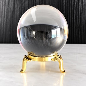 May include: A clear, spherical glass orb resting on a polished gold-toned metal stand. The stand features three curved legs and a circular base with a beaded edge. The glass sphere reflects the surrounding environment on its smooth surface.