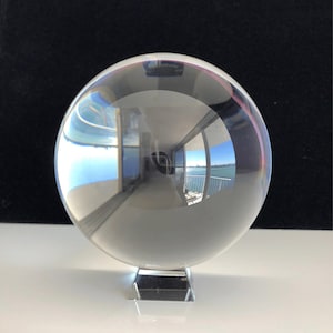 May include: A clear, spherical glass orb resting on a small, square, transparent glass base. The smooth, polished surface of the sphere creates a distorted, wide-angle reflection of a room with windows overlooking a body of water and a distant shore.