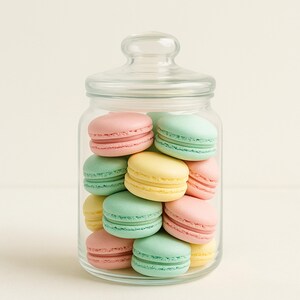 Pode incluir: Um frasco de vidro transparente cheio de macarons coloridos. Os macarons são em tons de rosa, amarelo e verde. O frasco tem uma tampa de vidro e está sobre uma superfície clara.
