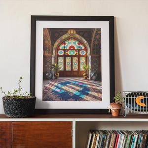 May include: A framed print of an ornate interior with a stained-glass window and wooden doors. The image features a colorful rug and architectural details. The print is displayed on a wooden cabinet with plants and vinyl records.