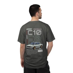 May include: Dark gray t-shirt featuring a graphic of a classic car with the text "HAKOSUKA" and "C10" in white. The design includes a checkered flag and additional text. The shirt is a short-sleeved crew neck.