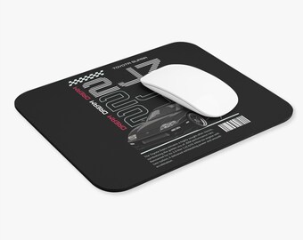 May include: Black rectangular mousepad featuring a graphic of a black Toyota Supra car. The mousepad has the text "JZA80" and "DREAM DREAM DREAM" in white and pink. A white computer mouse rests on the pad.