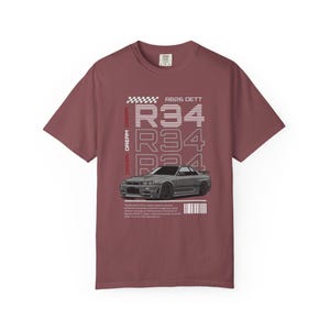 May include: A maroon t-shirt with a graphic of a gray sports car. The shirt features the text "R34" repeated vertically, along with "RB26 DETT" and other text. A checkered flag design is at the top.