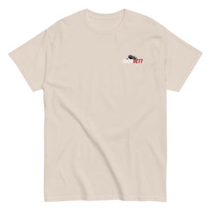 May include: A cream-coloured t-shirt with a crew neck. The shirt has a small logo on the upper left chest that reads "Raze Dett" in white and red. The t-shirt is made of a soft material.