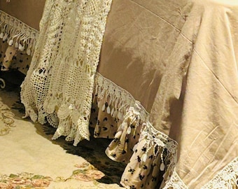 May include: A bed with a beige bedspread, adorned with decorative pillows in pink and checkered patterns. A cream-colored, open-weave lace runner drapes over the bed, complemented by a ruffled bed skirt with floral accents and tassel trim.
