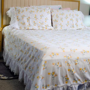 May include: A bed with a cream-colored headboard and a white floral-patterned bedspread. The bedding features yellow and orange flowers. The pillowcases and bed skirt have white lace trim, creating a vintage aesthetic.