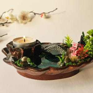 May include: A leaf-shaped candle holder with a miniature garden design. It features a small bridge, pink benches, and a lit candle. The holder is brown with green and blue details, creating a tranquil scene.