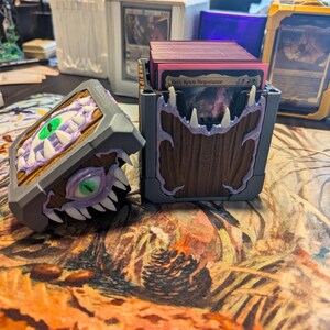 May include: Card storage boxes with a fantasy theme. The boxes are gray, brown, and purple, with a design of a monster's eye and teeth. The boxes are designed to hold trading cards. The boxes are on a patterned surface.