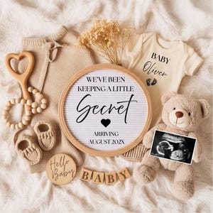 Könnte beinhalten: Eine Babyankündigung in neutralen Tönen mit einem Buchstabenbrett mit dem Text „We've been keeping a little secret“ und „Arriving August 202X“. Es gibt auch einen Teddybär, Babykleidung, einen Ultraschallscan und Holzblöcke mit dem Wort „BABY“.
