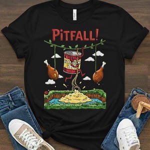May include: Black t-shirt with a humorous graphic featuring a can of beans labeled "Cranberry Sauce" swinging between two drumsticks, with the word "Pitfall!" above. The design is reminiscent of a video game.