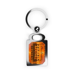 May include: A silver rectangular keyring with a circular loop. The rectangular pendant displays a group of silhouetted figures against an orange and yellow backdrop. The words "Bts Forever" are written in a script font.