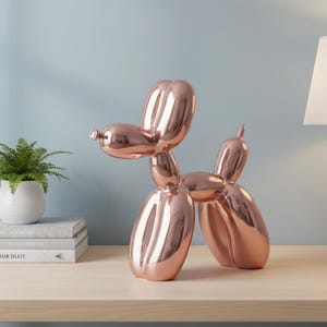 Könnte beinhalten: Eine roségoldene Ballonhund-Skulptur mit glänzender, reflektierender Oberfläche. Der Hund ist in einer stehenden Pose dargestellt, mit erhobenem Kopf und Schwanz. Die Skulptur steht auf einer hellen Holzoberfläche, mit einer Pflanze und Büchern im Hintergrund.