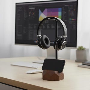 May include: A black and silver headset rests on a black metal stand, above a black smartphone and a geometric wooden base. The stand is on a light-colored wooden desk, with a computer monitor in the background.