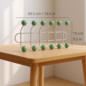 Könnte beinhalten: Ein metallener Wand-Kleiderhaken mit sechs grünen kugelförmigen Haken. Der Haken ist 49,5 cm breit und 15 cm hoch. Die Haken sind in einer Reihe angeordnet. Der Haken steht auf einer hellbraunen Holzoberfläche.