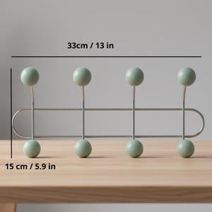 Può includere: Un attaccapanni da parete con quattro pioli sferici verde chiaro. Il portabiti è largo 33 cm e alto 15 cm. I pioli sono attaccati a una barra di metallo argentato con un design curvo.