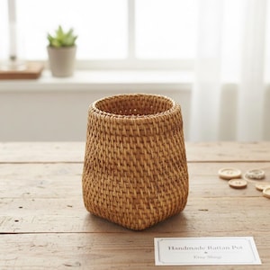 Op de afbeelding: Een handgemaakte rotan pot met een geweven ontwerp. De pot is lichtbruin van kleur en heeft een licht taps toelopende vorm. De pot staat op een houten oppervlak. De tekst "Handmade Rattan Pot" is zichtbaar.