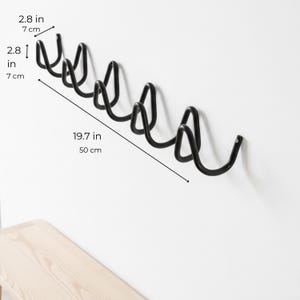 May include: A black metal wall-mounted coat rack with six curved hooks. The rack is 19.7 inches (50 cm) long and each hook is 2.8 inches (7 cm) high. The rack is mounted on a white wall.