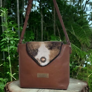May include: A brown leather crossbody bag with a flap closure featuring a cowhide panel in shades of brown, white, and black. The bag has a long, adjustable strap and a small metal plaque on the front. The bag is sitting on a tree stump.