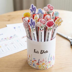 以下が含まれることがあります： 白い円筒形の容器に花柄のデザインとドイツ語のテキスト「Für Dich」があり、花の装飾が施されたペンが入っています。ペンにはさまざまな花のイラストと「bist die Beste」などのフレーズがあります。木製のテーブルには、はさみとスティックのりがあります。
