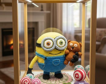 Peut inclure: Un personnage Minion au crochet, avec une peau jaune et une salopette bleue, tenant un petit ours en peluche marron, exposé dans une vitrine en bois. La vitrine contient également des bonbons et des fleurs au crochet. L'arrière-plan montre un salon avec une cheminée.