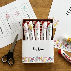 May include: A box of floral-themed gifts, including decorated pens, greeting cards, and bookmarks. The pens are white with floral designs and German text. The cards say "Happy Birthday" and "Danke". Scissors and glue sticks are also present.