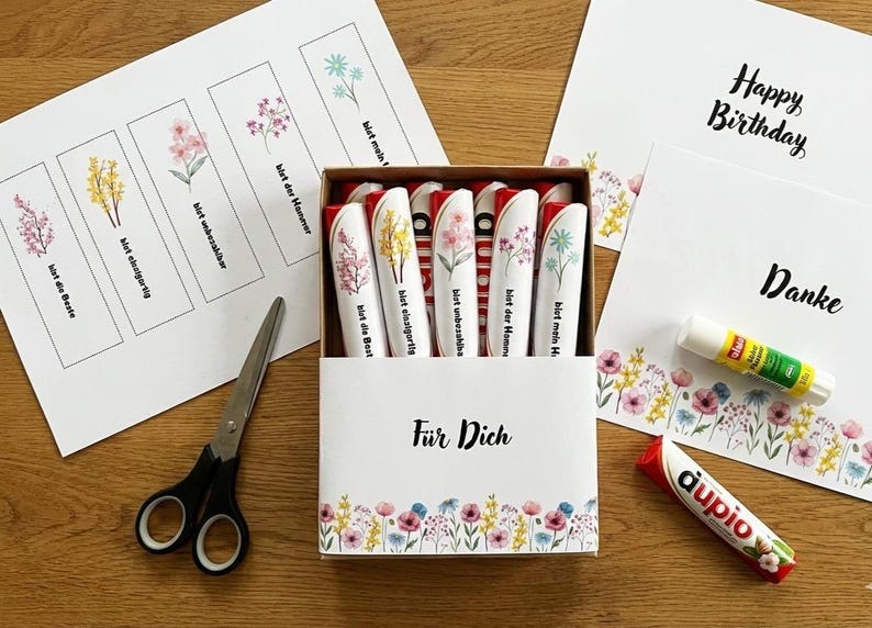May include: A box of floral-themed gifts, including decorated pens, greeting cards, and bookmarks. The pens are white with floral designs and German text. The cards say "Happy Birthday" and "Danke". Scissors and glue sticks are also present.