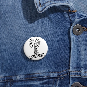 Peut inclure: Un badge blanc avec un dessin noir d'une tour radio et d'une antenne circulaire. Le texte "HAM RADIO" est imprimé en noir en dessous. Le badge est épinglé sur une veste en jean bleue.