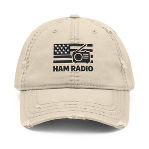 May include: A distressed beige baseball cap featuring a black graphic of an American flag with a radio and the text "HAM RADIO." The cap has a pre-worn look with frayed edges, suggesting a vintage or worn aesthetic. The design is centered on the front of the cap.
