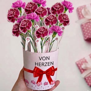 Op de afbeelding: Een papieren bloemstuk in een cilindrische container met een rode strik en de woorden "VON HERZEN". De bloemen zijn een mix van donkerroze en lichtroze anjers. De container heeft een bloemenpatroon aan de onderkant.