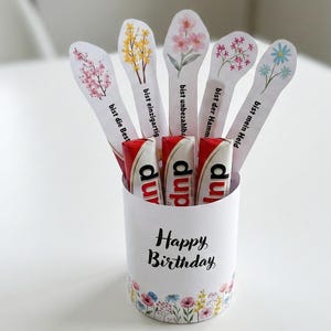 May include: A white cylindrical container with a floral design and the words "Happy Birthday" holds several candy bars and decorative floral-topped sticks. The sticks have German phrases.