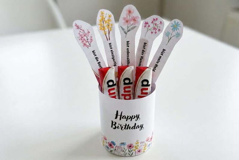 May include: A white cylindrical container with a floral design and the words "Happy Birthday" holds several candy bars and decorative floral-topped sticks. The sticks have German phrases.