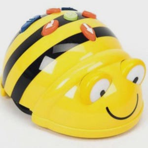 May include: A yellow and black bee-shaped robot toy with a rounded body. The top features several colourful control buttons, including orange arrows, a green button, and a blue button. The front has two white eyes and a black curved smile.