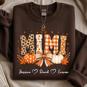 May include: A dark brown crewneck sweatshirt featuring the word MIMI in large, patterned block letters with leopard print, floral, orange gingham, and polka dot designs. Below the text are two pumpkins, a bow, and the names Jessica, David, and Emma.