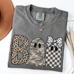 May include: A charcoal gray short-sleeve t-shirt featuring the word BOO in large letters. The B has a leopard print, the first O is a ghost with a camouflage pattern, and the second O is a ghost with a checkered pattern and a white polka dot bow.