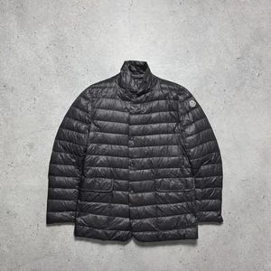 Puede incluir: Chaqueta acolchada negra con cuello alto y cierre de botones. La chaqueta tiene dos bolsillos delanteros y un pequeño parche con el logotipo en la manga superior izquierda. Diseñada para calidez y estilo.