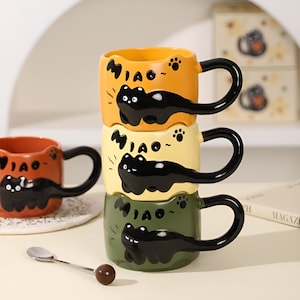 May include: Three stacked ceramic mugs in shades of orange, yellow, and green, each adorned with a black cat design and the word "Miao." The mugs have black handles and paw prints. A small spoon with a brown ball is in front.