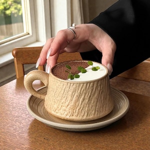 May include: A beige ceramic mug with a textured finish, containing a creamy drink, dusted with cocoa and garnished with green leaves. The mug rests on a matching saucer. The handle is a rounded shape.