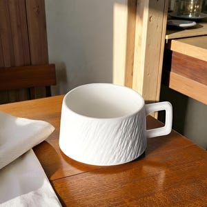 May include: A white ceramic mug with a textured, bark-like pattern. The mug has a square handle. It is on a wooden table. The mug is empty. The mug is ready to be filled with a beverage.
