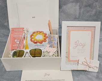 Peut inclure: Une boîte cadeau blanche remplie d'articles, dont des lunettes de soleil, des collations et un stylo. Une carte encadrée avec le mot "Joy" et une petite étiquette cadeau sont également présentes. La boîte et le cadre portent le mot "Joy".