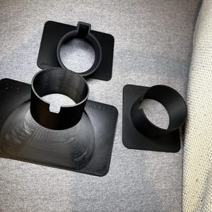 May include: Three black, 3D-printed plastic components. One is a circular ring attached to a rectangular base. Another is a smaller ring with a rectangular base. The third is a larger, flared cylinder with a rectangular base. The items are on a grey surface.