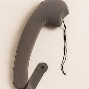 May include: A gray neck support pillow with a curved design. The pillow has a rounded top and a strap with a button closure. A black string is attached to the top. The pillow is made of a soft, flexible material.