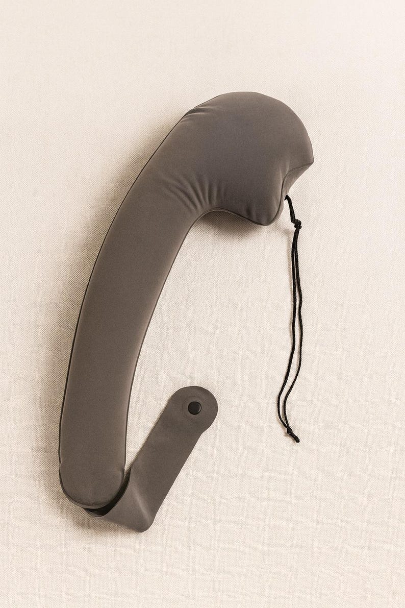 May include: A gray neck support pillow with a curved design. The pillow has a rounded top and a strap with a button closure. A black string is attached to the top. The pillow is made of a soft, flexible material.