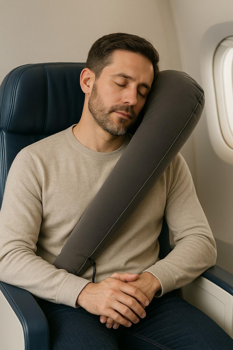 May include: A gray travel pillow is wrapped around the neck of a person seated on an airplane. The person is wearing a beige sweater and is resting their head on the pillow. The airplane seat is blue.