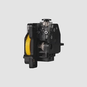 May include: A black mechanical component featuring a prominent yellow gear on the left side, a central metallic knurled wheel, and a small clear plastic housing at the top. The assembly includes various mounting points and internal electronic parts.